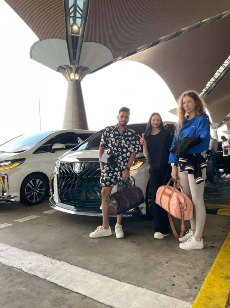 Three travellers with bags beside a white MPV at an airport pickup bay